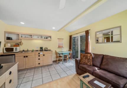 A small kitchen and dining area with light wood cabinets, a microwave, and a coffee maker. A brown sofa and glass table with two chairs sit near a window and door leading outside. The walls are painted yellow.