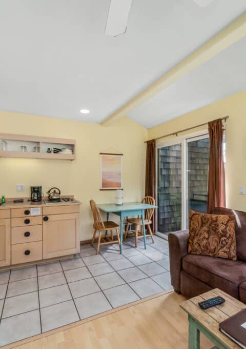 A small kitchen and dining area with light wood cabinets, a microwave, and a coffee maker. A brown sofa and glass table with two chairs sit near a window and door leading outside. The walls are painted yellow.