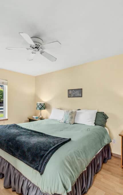 A bedroom with a large bed, two patterned lamps on nightstands, a black dresser, light wood floors, a ceiling fan, and a window showing greenery and a parked car outside.