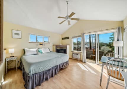 A bright bedroom with a king-size bed, fireplace, small table and chairs, ceiling fan, large windows, and sliding glass doors leading to a balcony. Light wood flooring and pale walls add to the airy feel.