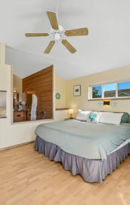 A bedroom with a king-size bed, light wood flooring, a fireplace, a ceiling fan, a small desk, and a partial dividing wall with a view into a bathroom. A window lets in natural light.