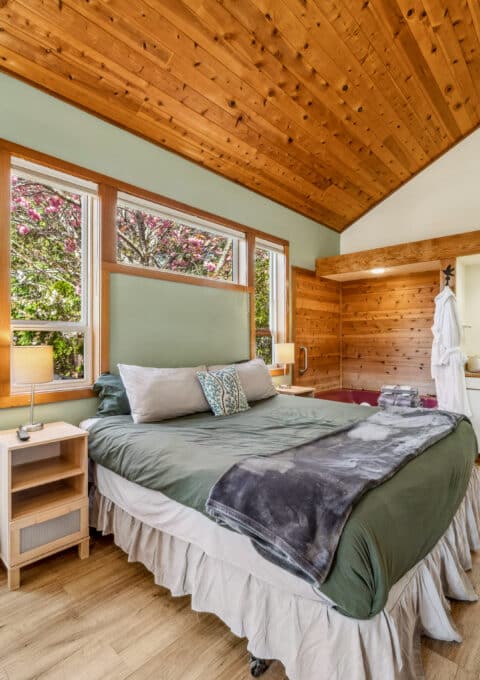 A bedroom with a green bedspread, wood-paneled ceiling, and light wood floors. There is a kitchenette, a door leading outside, large windows, and a robe hanging by the sink. Natural light fills the room.