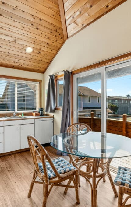 A small kitchen and dining area with white cabinets, wood ceiling, a round glass table with three wicker chairs, and large windows offering a view of houses outside.