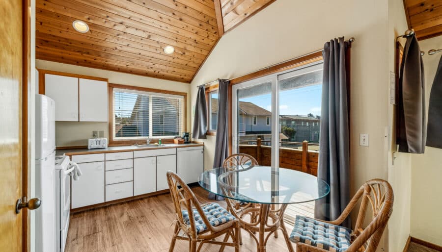 A small kitchen and dining area with white cabinets, wood ceiling, a round glass table with three wicker chairs, and large windows offering a view of houses outside.