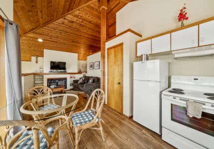 A cozy kitchen and living area with wood-paneled ceiling, white appliances, a round glass dining table with four wicker chairs, a gray sofa, wall-mounted TV, and a fireplace. Natural light comes through a window.