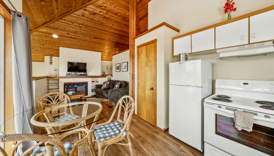 A cozy kitchen and living area with wood-paneled ceiling, white appliances, a round glass dining table with four wicker chairs, a gray sofa, wall-mounted TV, and a fireplace. Natural light comes through a window.