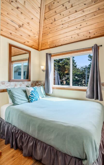 A small bedroom with a bed covered in light blue bedding, a wood-paneled ceiling, light-colored walls with decorative trim, a window with gray curtains, a wall mirror, and a closed wooden door.