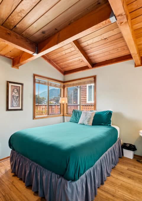A small bedroom with a wooden ceiling, a double bed with teal bedding, two wall lamps, a bedside table with a lamp, framed art on the wall, and a window overlooking an outdoor scene.