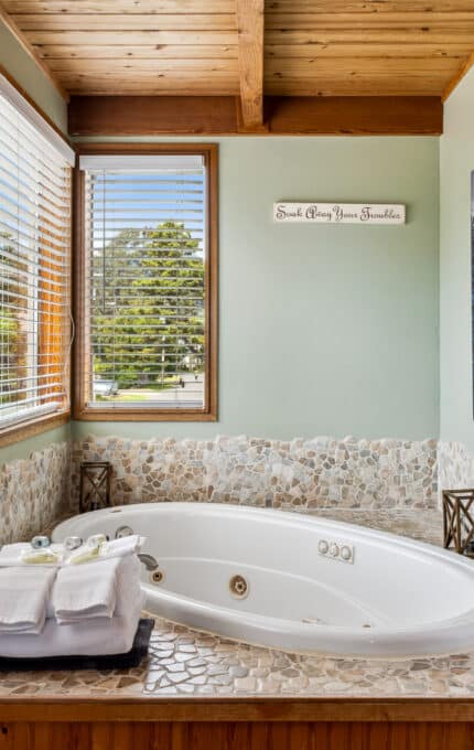 A corner bathtub with stone tile surround sits beneath two large windows with blinds. Towels, toiletries, and a white bathrobe hang nearby. A wave painting and a decorative sign are on the pale green walls.