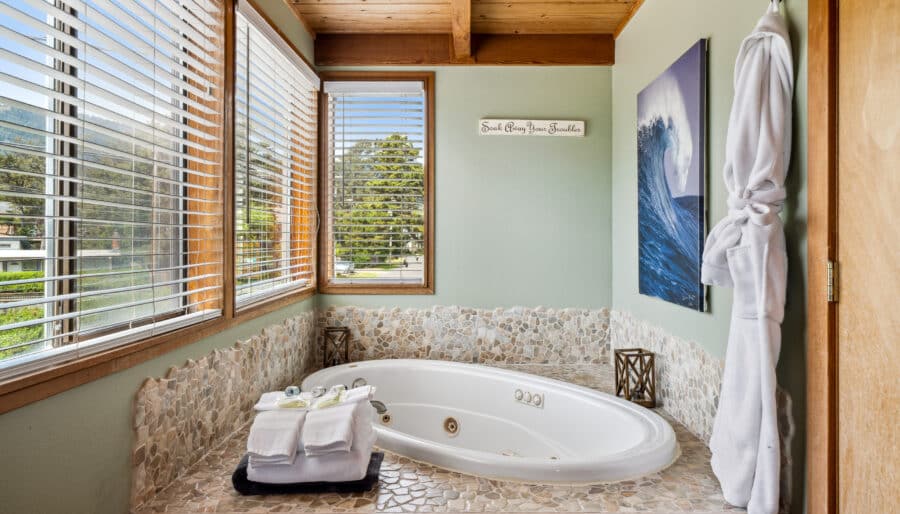 A corner bathtub with stone tile surround sits beneath two large windows with blinds. Towels, toiletries, and a white bathrobe hang nearby. A wave painting and a decorative sign are on the pale green walls.