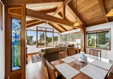 A dining room with a wooden table and chairs opens to a living area with couches. Large windows provide natural light, and a stained glass panel decorates the left side. Exposed wood beams are visible on the ceiling.