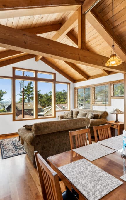 A dining room with a wooden table and chairs opens to a living area with couches. Large windows provide natural light, and a stained glass panel decorates the left side. Exposed wood beams are visible on the ceiling.