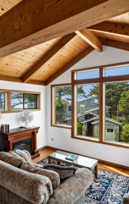 A living room with wood-paneled vaulted ceilings, large windows, two sofas, a patterned rug, a coffee table, a lamp, a fan, and a fireplace. Trees and neighboring houses are visible outside.