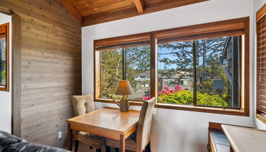 A small dining table with two upholstered chairs is placed by large windows, overlooking a garden with colorful flowers and trees. A lamp sits on the table in a room with wood-paneled walls and a sloped wooden ceiling.