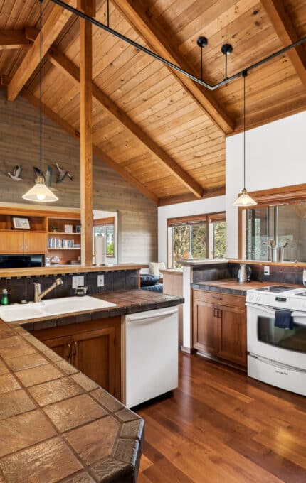 Spacious kitchen with wood-paneled walls and ceiling, brown tile countertops, white appliances, and large windows. There are hanging lights and wooden flooring throughout the room.