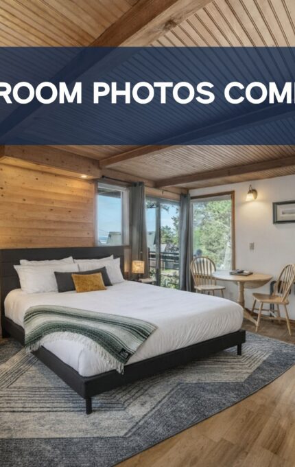 A modern hotel room with wood-paneled walls and ceiling, a bed with white linens, a small dining table, a fireplace, a flat-screen TV, and large windows. Banner text reads "UPDATED ROOM PHOTOS COMING SOON!.