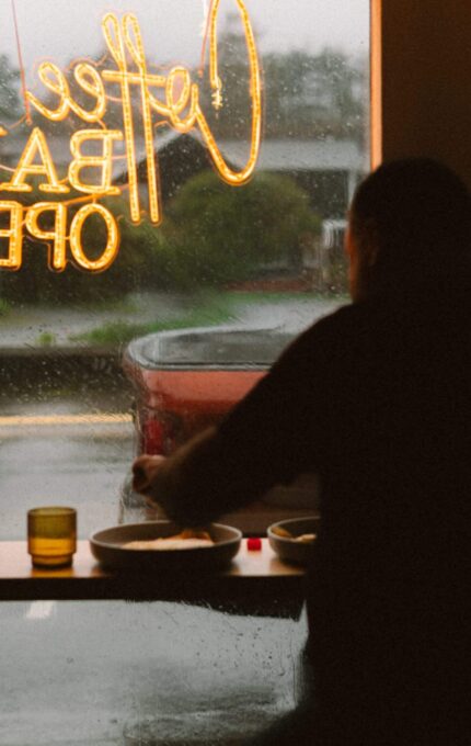 Cosy hotel café with large window, warm lighting, guest dining, and view of rainy street enhancing relaxed atmosphere.