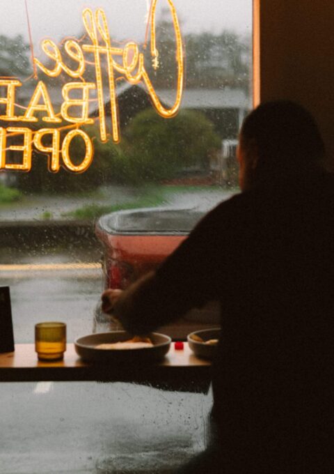 Cosy hotel café with large window, warm lighting, guest dining, and view of rainy street enhancing relaxed atmosphere.