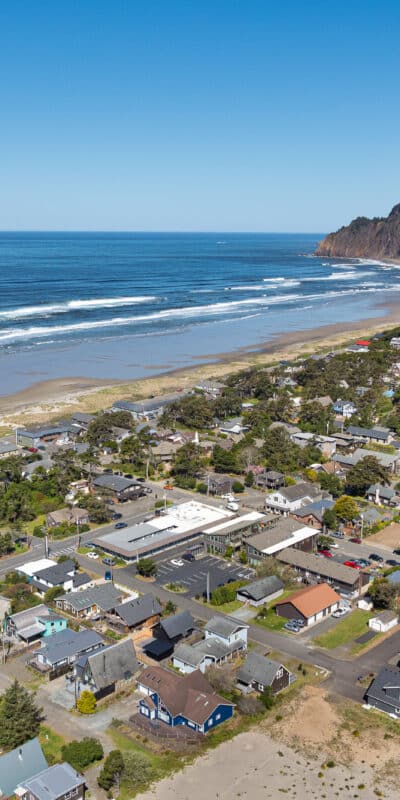 Aerial view of a coastal town with houses, roads, and trees next to a sandy beach and ocean. A forested hill rises in the background under a clear blue sky.