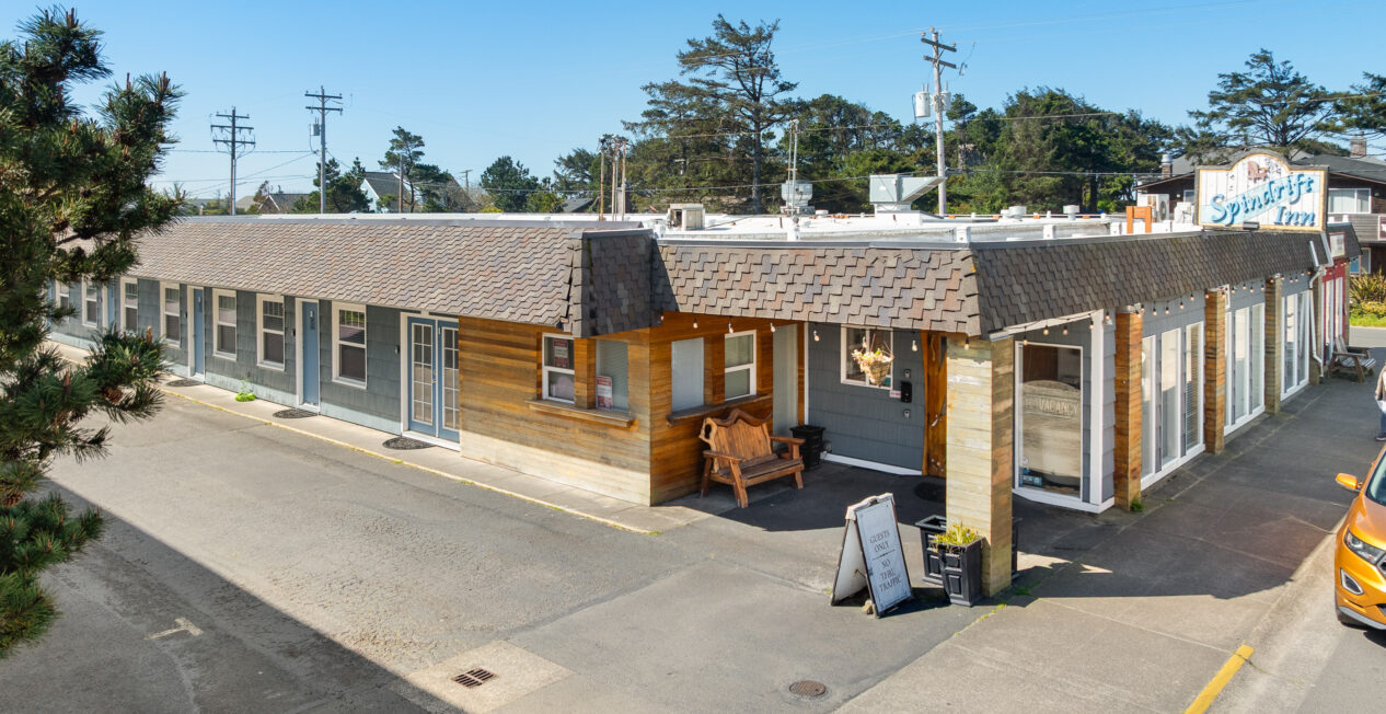 Single-story motel building with a shingled roof, a wooden porch area with a bench, and a sign reading "Spindrift Inn" on the roof. A yellow car is parked nearby under a clear blue sky.