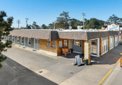 Single-story motel building with a shingled roof, a wooden porch area with a bench, and a sign reading "Spindrift Inn" on the roof. A yellow car is parked nearby under a clear blue sky.