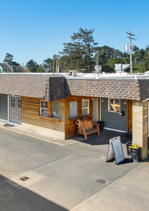Single-story motel building with a shingled roof, a wooden porch area with a bench, and a sign reading "Spindrift Inn" on the roof. A yellow car is parked nearby under a clear blue sky.