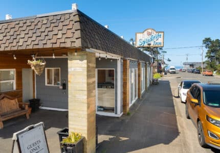 A small hotel with a shingled roof and wooden pillars sits next to a sidewalk. A sign reads “Sea Spirit Inn.” Several cars are parked along the street, and a bench and hanging basket are near the entrance.