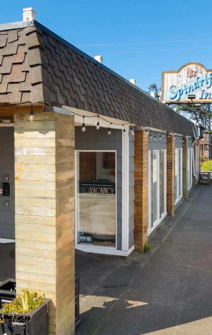 A small hotel with a shingled roof and wooden pillars sits next to a sidewalk. A sign reads “Sea Spirit Inn.” Several cars are parked along the street, and a bench and hanging basket are near the entrance.