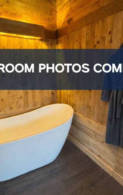 A modern bathroom with wood-paneled walls, a white freestanding bathtub, two gray robes hanging on hooks, and a blue banner reading "UPDATED ROOM PHOTOS COMING SOON!" across the center.