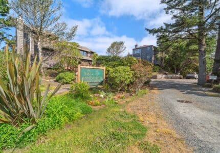 A gravel driveway leads to a wooden sign reading "The Inn at Manzanita," surrounded by greenery and flowers, with several buildings and parked cars visible in the background under a partly cloudy sky.