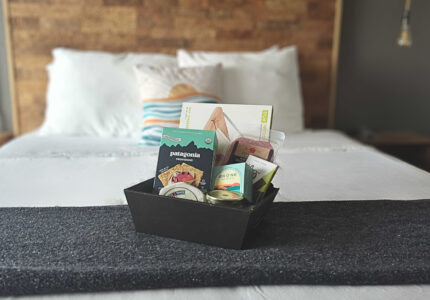 A black rectangular gift basket containing snacks and small items sits on a folded gray blanket at the foot of a neatly made white bed with pillows in a softly lit room.