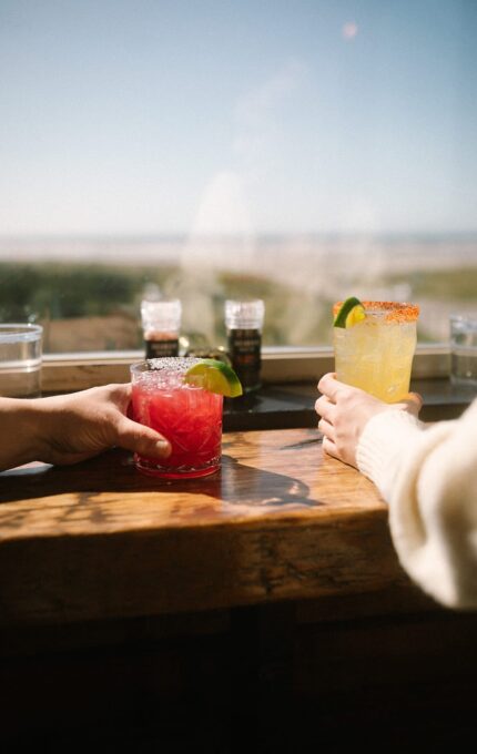 Two people holding cocktails in short glasses on a wooden table near a window. One drink is red with a lime wedge, and the other is yellow with a chili rim and lime. Daylight and an outdoor view are visible in the background.