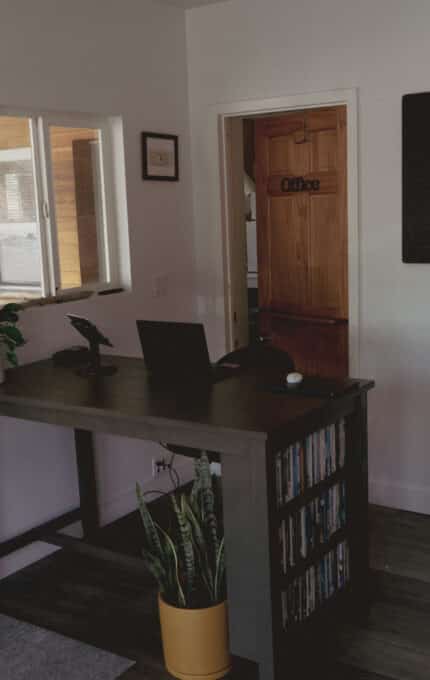 A small office entry area with a wooden door, a tall desk with a laptop, a yellow potted plant, and two letter board signs on the wall. A record player sits on a side table near a wooden door labeled “Office.”.