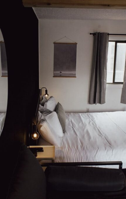 A neatly made bed with white linens is positioned against a white wall between two windows with gray curtains. A framed picture hangs above the bed. The room is reflected in a round mirror on the left.