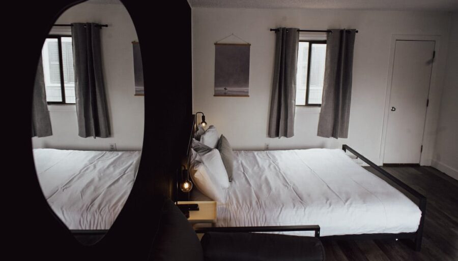 A neatly made bed with white linens is positioned against a white wall between two windows with gray curtains. A framed picture hangs above the bed. The room is reflected in a round mirror on the left.