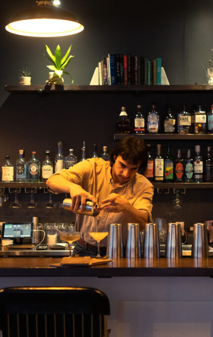 A bartender stands behind a well-stocked bar, pouring liquid into a shaker. Shelves with bottles, glasses, books, and decorations are visible behind him. The bar is lit by three overhead lights.