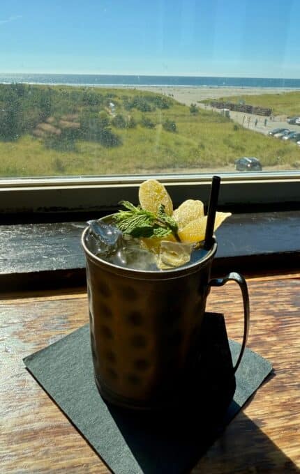 A copper mug filled with an iced drink garnished with mint and candied ginger sits on a black napkin by a window, with a view of grassy dunes, parked cars, and the ocean under a clear, sunny sky.