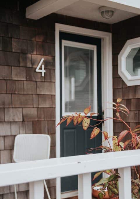 A dark blue door with a window and white trim is set in a shingle-clad wall with the number 4 displayed beside it. A white chair and some leafy plants are visible in the foreground behind a white railing.