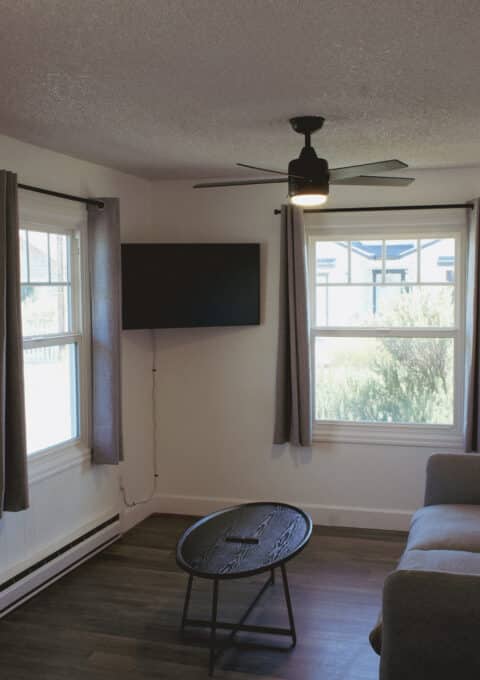 A small living room with a gray sofa, round black coffee table, wall-mounted TV, ceiling fan, two windows with gray curtains, a wall light, and a white door. The room has wood flooring and white walls.