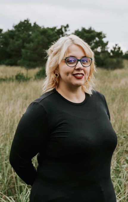 A person with blonde hair, glasses, and a black long-sleeve top stands in a grassy field with trees in the background on a cloudy day.