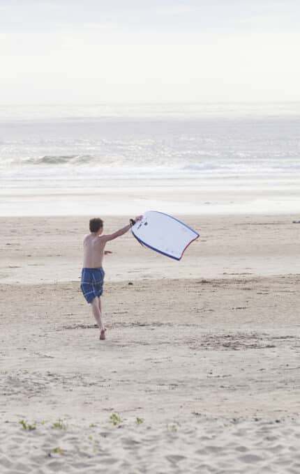 Two boys in blue swim trunks run on a sandy beach toward the ocean, each carrying a boogie board. The sky is overcast, and the beach is mostly empty with calm waves in the background.