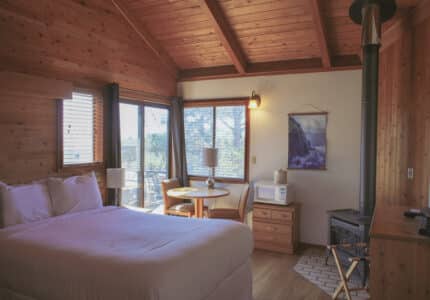 A cozy cabin room with wood-paneled walls and ceiling, a queen bed, small dining table with two chairs, microwave, dresser, wood stove, and large windows letting in natural light.