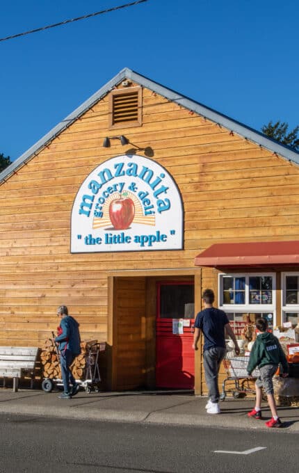 A small group of people stand outside Manzanita Grocery & Deli, a wooden building with a sign reading "the little apple." Some are pushing shopping carts near outdoor produce bins. The sky is clear and blue.