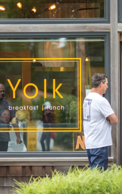 Several people stand and talk outside the entrance of a restaurant with large windows. The window features a yellow sign that reads "Yolk breakfast lunch." The surroundings include greenery and a wooden building exterior.