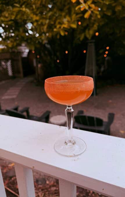A coupe glass filled with an orange cocktail, topped with a spiced or sugar rim, sits on a white railing. In the background, there are trees, string lights, and outdoor seating.