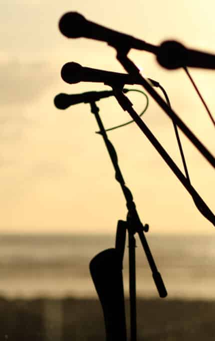 Four microphone stands are silhouetted against a beach sunset, with the ocean and sky in the blurred background. The image is backlit, emphasizing the outlines of the microphones and stands.