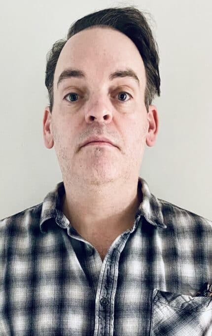 A man with short brown hair and a neutral expression is wearing a black-and-white plaid button-up shirt and standing against a plain off-white background.