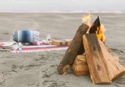 A small campfire burns on a sandy beach near a neatly arranged blanket, a rolled-up throw, and several items including snacks and a cardboard box. The ocean is visible in the background under cloudy skies.