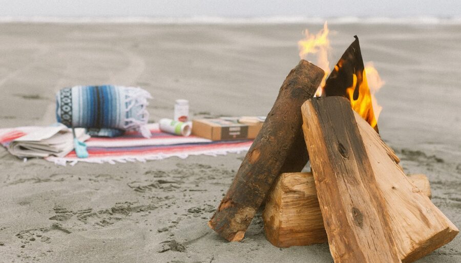 A small campfire burns on a sandy beach near a neatly arranged blanket, a rolled-up throw, and several items including snacks and a cardboard box. The ocean is visible in the background under cloudy skies.