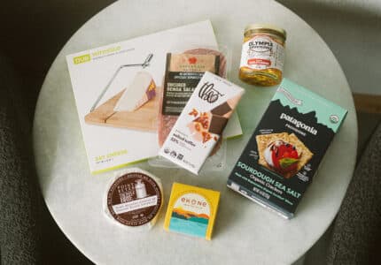 A round table displays a cheese slicer, uncured salami, sea salt crispbread, a jar of marinated olives, chocolate, smoked salmon, and a round tin of cheese, all in their packaging.